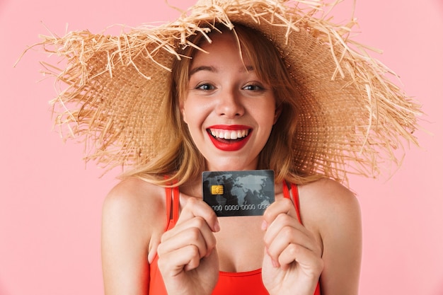 Retrato de mujer joven complacida con pelo largo y rizado con sombrero de paja de verano sonriendo y sosteniendo una tarjeta de crédito aislada sobre pared rosa