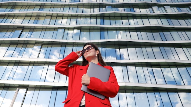 Retrato de una mujer joven con una computadora portátil en el fondo de un edificio Un oficinista en una chaqueta roja Freelancer afuera