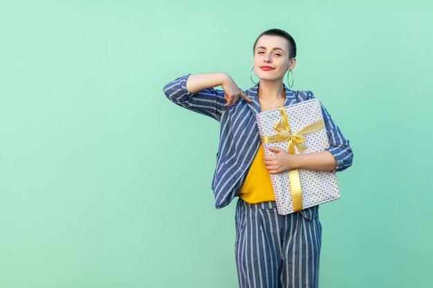 Retrato de mujer joven confiada hermosa con pelo corto en traje a rayas de pie, señalando con el dedo a sí misma y sosteniendo la caja actual con rostro orgulloso. Interior, aislado, foto de estudio, fondo verde