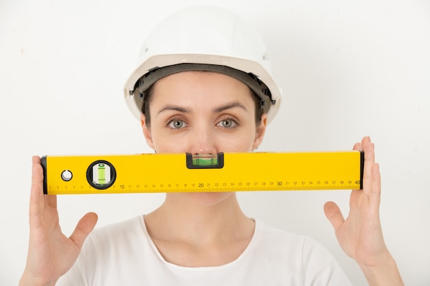 Foto retrato de mujer joven confiada positiva ajustando el casco y sosteniendo el rollo de pintura contra la pared blanca