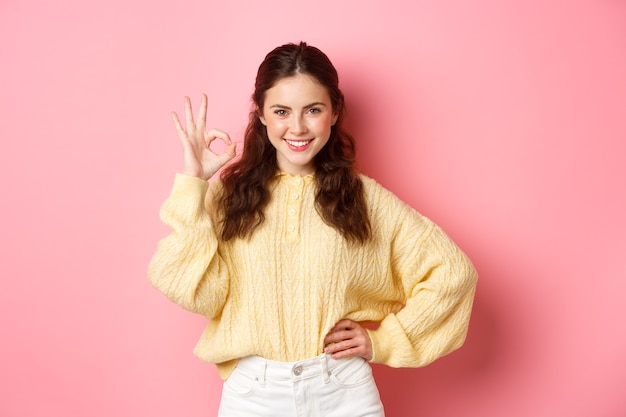 Retrato de mujer joven confiada sonriendo complacida, diga que sí mientras muestra un signo de bien, dé su aprobación, alabe la excelente elección, de pie contra la pared rosa.