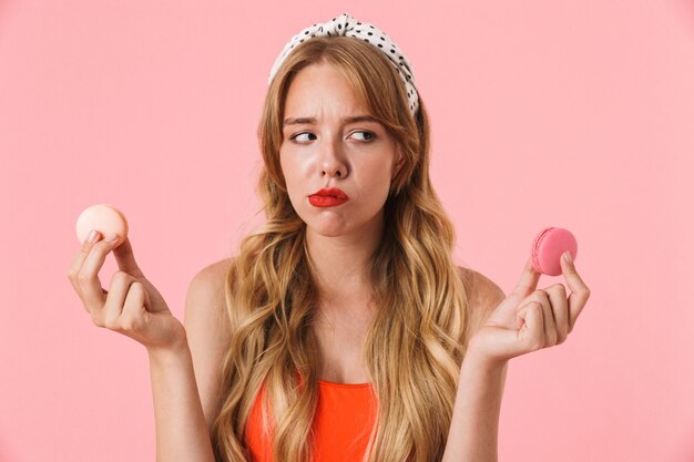 Retrato de mujer joven disgustada con pelo largo y rizado dudando mientras sostiene galletas macaron aislado sobre pared rosa