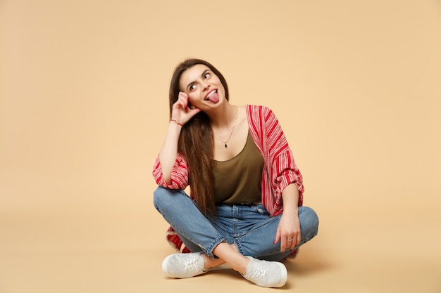 Retrato de mujer joven divertida loca en ropa casual sentada, mirando a un lado, mostrando la lengua aislada sobre fondo de pared beige pastel. Personas sinceras emociones, concepto de estilo de vida. Simulacros de espacio de copia.