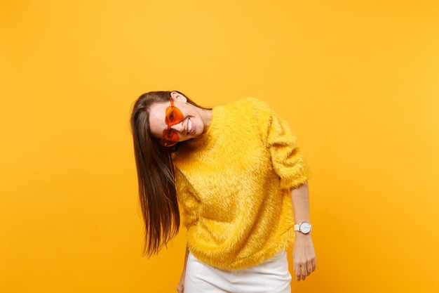 Retrato de mujer joven divertida sonriente alegre en suéter de piel, pantalón blanco y anteojos naranja corazón aislado sobre fondo amarillo brillante. Personas sinceras emociones, concepto de estilo de vida. Área de publicidad.