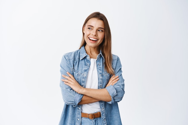 Retrato de mujer joven elegante con cabello rubio, brazos cruzados sobre el pecho, mirando a la izquierda en el espacio vacío para pancarta y sonriendo complacido, mirando publicidad, pared blanca