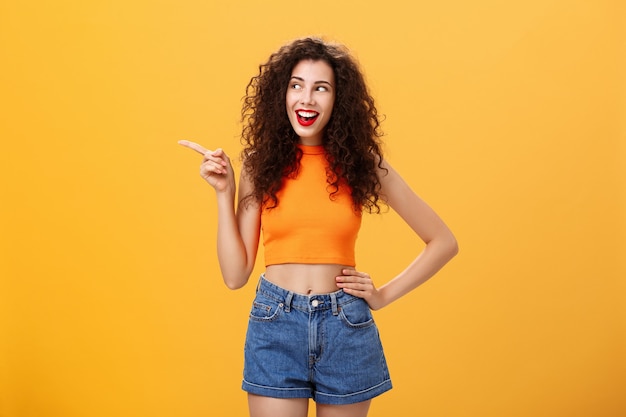 Retrato de mujer joven elegante y entusiasta encantada con peinado rizado en lápiz labial rojo y parte superior recortada sosteniendo la mano en la cintura apuntando y mirando a la izquierda divertido y feliz sobre fondo naranja.