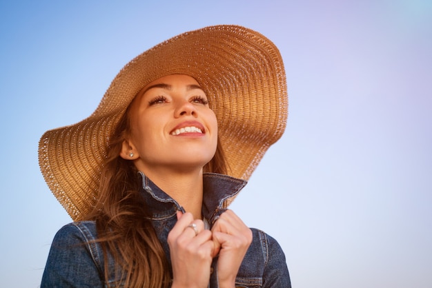 Retrato de una mujer joven elegante feliz en una chaqueta de mezclilla blanca y un sombrero de paja en la orilla del océano al atardecer, mirando a lo lejos