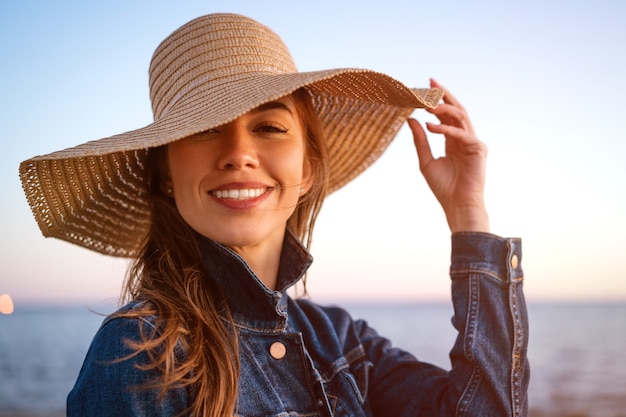 Retrato de una mujer joven elegante feliz en una chaqueta de mezclilla blanca y un sombrero de paja en la orilla del océano al atardecer, mirando a lo lejos