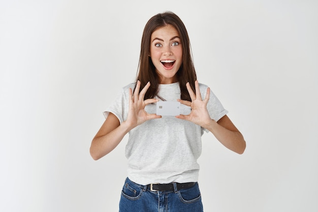 Retrato de una mujer joven emocionada vestida con camiseta y jeans, sosteniendo una tarjeta de crédito y sonriendo al frente, pared blanca