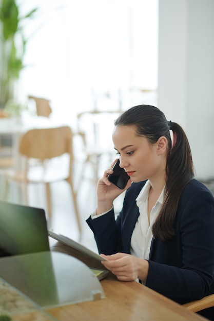 Retrato de mujer joven empresaria seria hablando por teléfono y leyendo un documento sobre el desarrollo empresarial