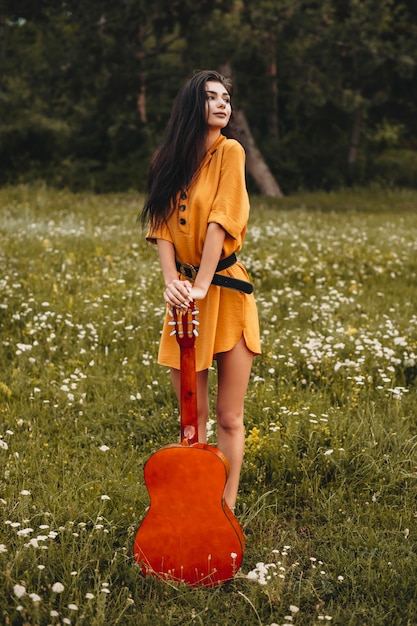 Retrato de una mujer joven encantadora, apoyado en una guitarra y apartar la mirada al aire libre. Joven freelance haciendo contenido para su canal en un campo y con una guitarra.