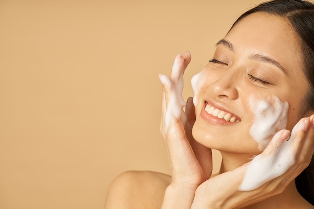 Retrato de mujer joven encantadora sonriendo con los ojos cerrados mientras aplica un limpiador facial de espuma suave.