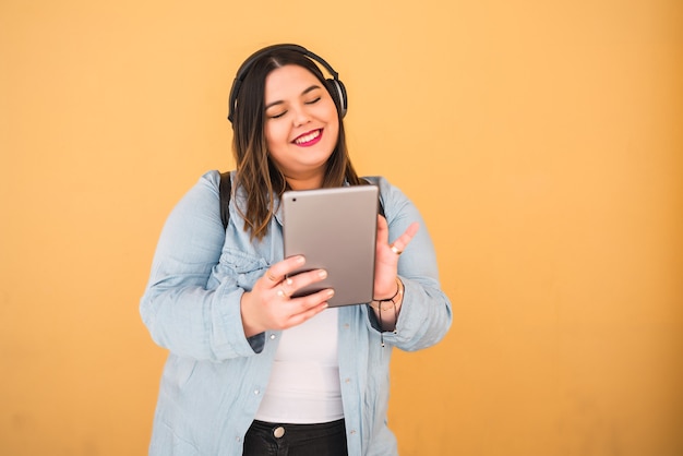 Retrato de mujer joven escuchando música con auriculares y tableta digital al aire libre contra la pared amarilla