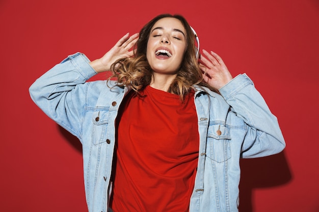 Retrato de una mujer joven con estilo alegre vistiendo chaqueta de mezclilla que se encuentran aisladas sobre la pared roja, escuchando música con auriculares, bailando