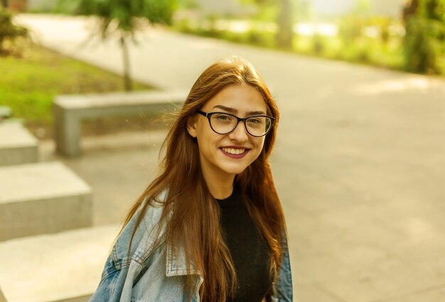 Retrato de una mujer joven estudiante en una chaqueta vaquera y gafas en un parque al aire libre