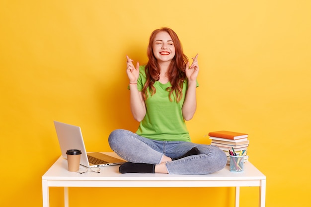 Retrato de mujer joven estudiante sentada en su escritorio y cruzando los dedos, desea tener suerte para proyectos futuros, tiene expresión emocionada, cierra los ojos, usa casualmente, aislado sobre una pared amarilla.