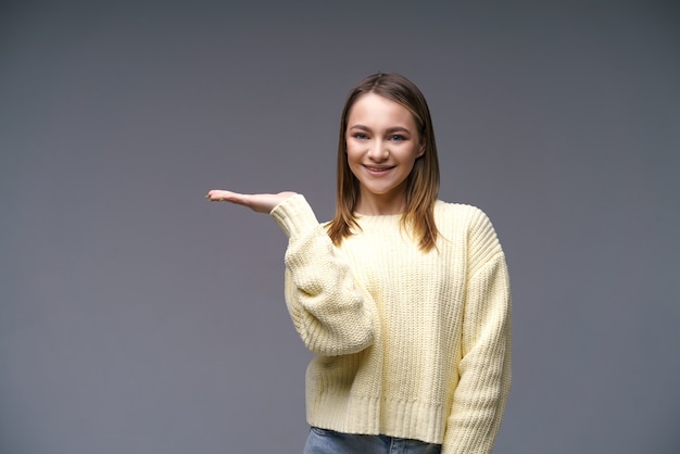 Retrato de una mujer joven de etnia caucásica muestra con la mano hacia un lado en un suéter amarillo ...