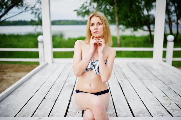 Retrato de una mujer joven fabulosa en bikini sentada y posando en un mirador de madera blanca en el parque al lado del lago