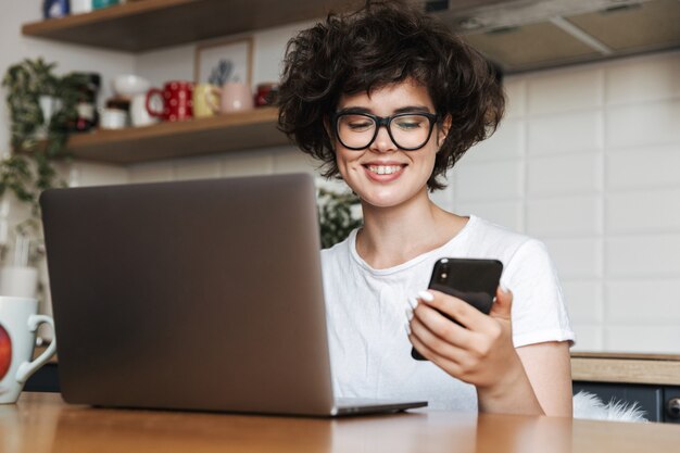 Retrato de una mujer joven feliz con anteojos trabajando en la computadora portátil en casa por la mañana, sosteniendo el teléfono móvil