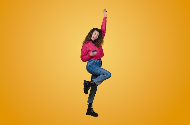 Foto retrato de una mujer joven feliz celebrando la victoria sosteniendo un puño mientras sostiene el teléfono móvil aislado sobre fondo amarillo gente feliz emoción positiva felicidad ganar éxito