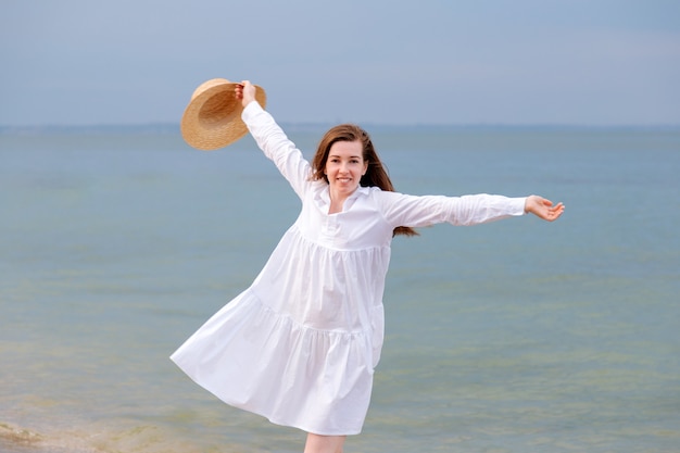 Retrato de mujer joven feliz disfrutando de vestido blanco que descansan al atardecer en la playa del mar. Mujer feliz con sombrero de paja en la mano en la orilla del mar.