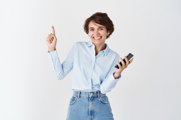 Retrato de mujer joven feliz escuchando música en auriculares inalámbricos, sosteniendo el teléfono móvil y apuntando hacia arriba, pared blanca