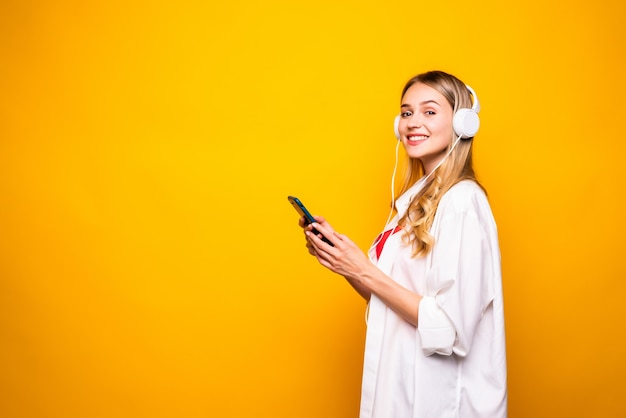 Retrato de una mujer joven feliz escuchando música con auriculares y teléfono móvil aislado sobre pared amarilla