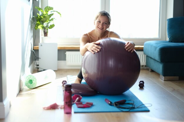 Retrato de mujer joven feliz con gran pelota de gimnasia sobre una alfombra en casa. Ejercicios de fitball en concepto de casa