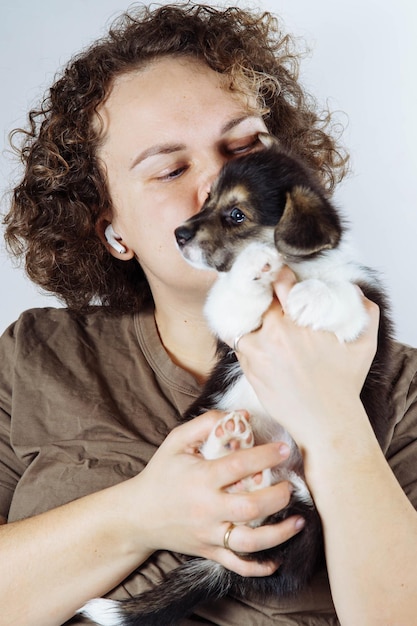 Foto retrato de una mujer joven feliz con el pelo rizado vistiendo una camiseta marrón sosteniendo las manos besando a un pequeño cachorro blanco y negro de perro corgi pembroke galés sobre fondo gris amor de mascotas cuidado de mascotas vertical