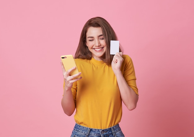 Retrato de una mujer joven feliz que sostiene el teléfono móvil y la tarjeta de crédito aislados sobre rosa.