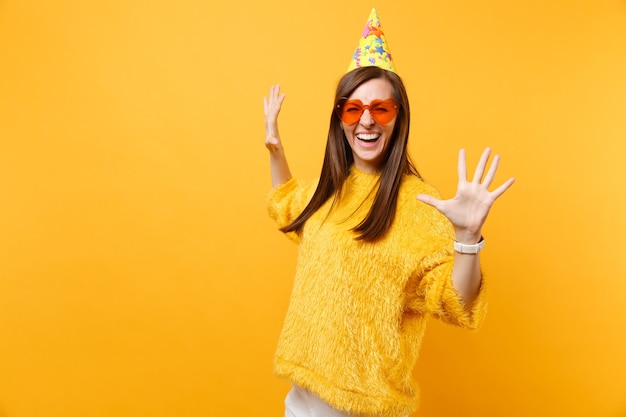 Retrato de mujer joven feliz riendo en vasos de corazón naranja, sombrero de fiesta de cumpleaños extendiendo las manos aisladas sobre fondo amarillo brillante. Personas sinceras emociones, concepto de estilo de vida. Área de publicidad.