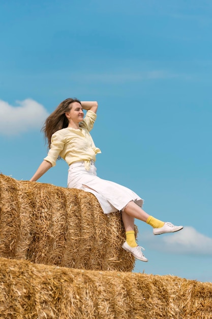 Retrato de mujer joven feliz sentada en un pajar alto sobre fondo de cielo azul Mujer descansando en el campo Cosecha