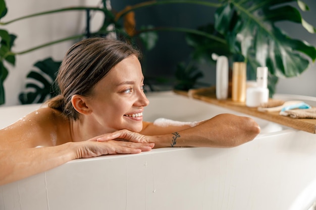 Retrato de mujer joven feliz sonriendo a un lado mientras descansa en la bañera en el spa de lujo