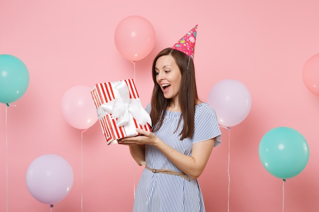 Retrato de mujer joven feliz sorprendida en vestido azul de sombrero de cumpleaños mantenga caja roja con regalo presente sobre fondo rosa pastel con coloridos globos de aire. Fiesta de cumpleaños, personas sinceras emociones.