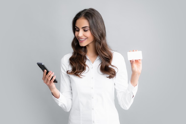 Retrato de una mujer joven feliz con tarjeta de crédito de plástico usando un teléfono móvil aislado sobre gris