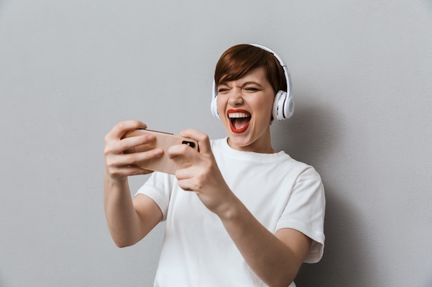 Retrato de mujer joven feliz usando audífonos jugando videojuegos en celular aislado sobre pared gris