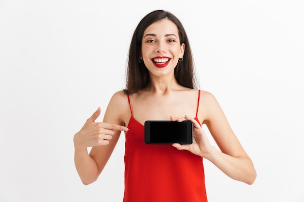 Retrato de una mujer joven feliz en vestido aislado, apuntando al teléfono móvil de pantalla en blanco