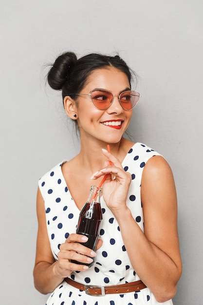 Retrato de una mujer joven feliz en vestido de verano y gafas de sol aisladas, sosteniendo la botella con bebida gaseosa