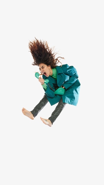 Retrato de mujer joven en gabardina verde saltando sobre fondo blanco gritando en megáfono en