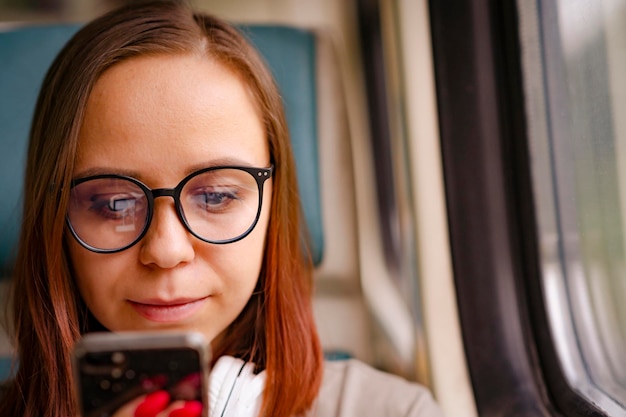 Retrato de una mujer joven con gafas navegando por un teléfono móvil en un tren Cerrar una mujer bonita usando un aparato mientras está sentada en un tren suburbano