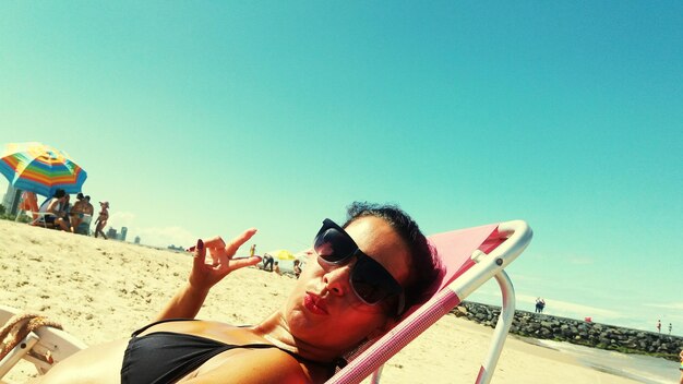 Foto retrato de una mujer joven con gafas de sol relajándose en una silla de sol en la playa contra el cielo azul