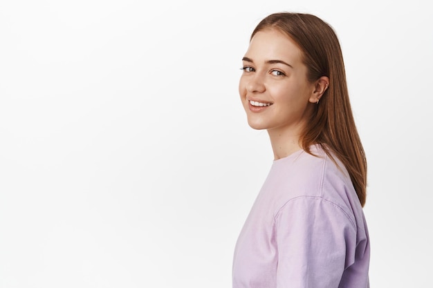 Retrato de mujer joven girando la cabeza a la cámara, sonriendo feliz, alegre expresión de la cara, de pie en camiseta púrpura contra fondo blanco.