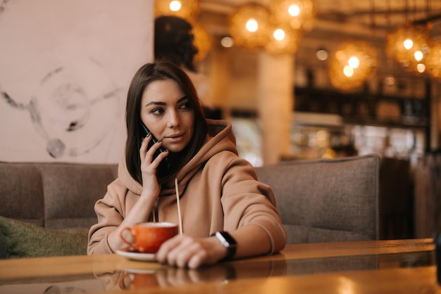 Retrato de una mujer joven y guapa con ropa informal hablando por teléfono en una mesa en un café acogedor Taza de café sobre la mesa Concepto de actividad de ocio