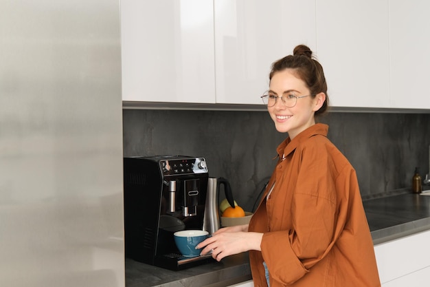 Retrato de una mujer joven haciéndose café de pie cerca de la máquina y sonriendo sosteniendo una taza para