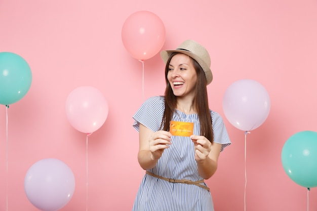 Retrato de mujer joven hermosa alegre en vestido azul de sombrero de paja de verano con tarjeta de crédito mirando a un lado sobre fondo rosa con globos de colores. fiesta de cumpleaños personas emociones sinceras.