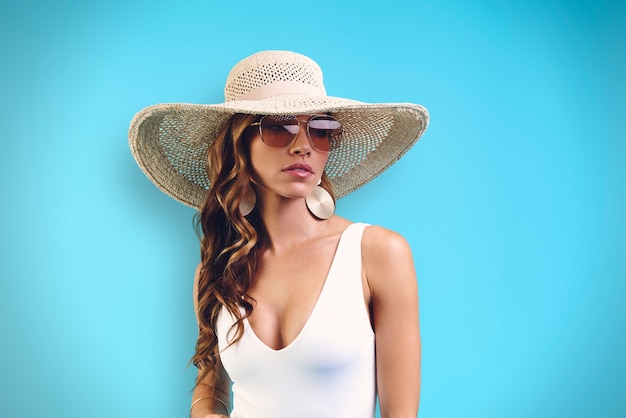 Retrato de mujer joven hermosa en elegante sombrero mirando a otro lado mientras está de pie contra el fondo azul.