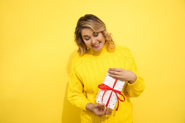 Retrato de mujer joven hermosa, exitosa, feliz y sonriente sobre fondo amarillo con caja de regalo en las manos, atada con cinta roja. Emociones de felicidad. Interesado en disfrutar de las vacaciones.