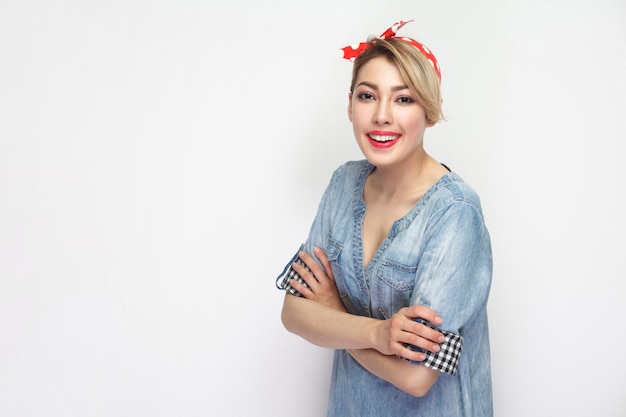 Retrato de mujer joven hermosa feliz satisfecha en camisa de mezclilla azul, maquillaje, diadema roja de pie con los brazos cruzados y mirando a la cámara con una gran sonrisa. Foto de estudio aislado sobre fondo blanco.