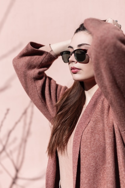 Retrato de mujer joven hermosa en gafas de sol de moda en abrigo al aire libre junto a la pared rosa en un día soleado. Hermosa chica con piel limpia con labios sensuales endereza el pelo lujoso de pie cerca del edificio vintage.