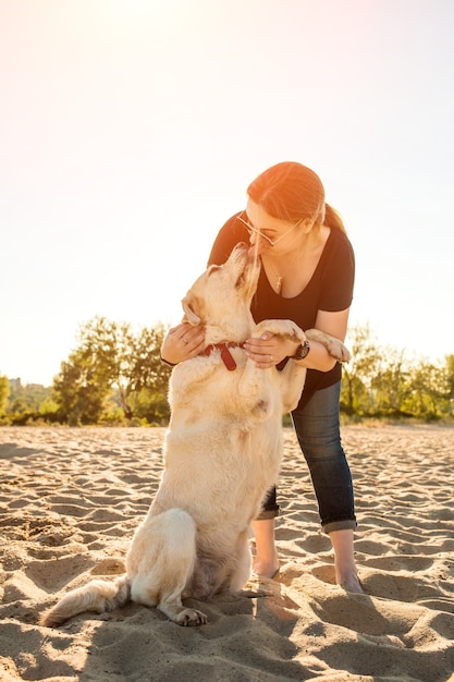 Retrato de mujer joven hermosa en gafas de sol sentado en la playa de arena con la niña de perro golden retriever ...